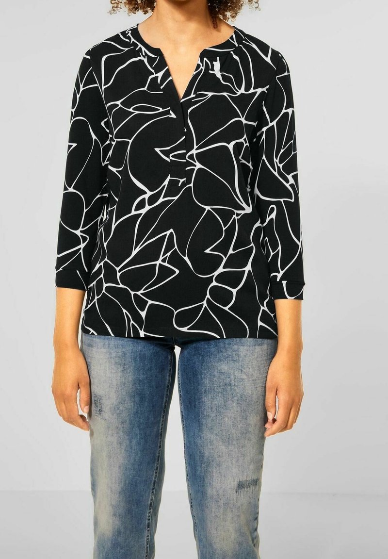 Svart topp med vita abstrakta linjemönster, trekvartsärmar och notch-krage. Buren med slitna blå jeans.