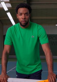 Grønn t-skjorte med korte ermer og et lite Lacoste-logo, sammen med mørke shorts, båret av en person som står på en tennisbane.