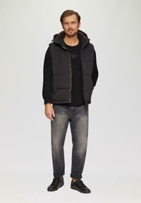 Zwarte gewatteerde vest met een ritssluiting en drukknoopsluiting, gedragen over een zwart T-shirt. Grijze spijkerbroek en zwarte sneakers maken de outfit compleet.