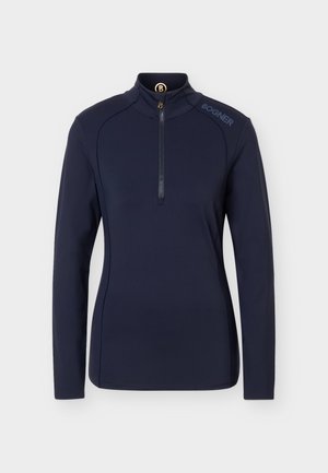 Persona de pie, mirando hacia adelante, vistiendo una camiseta negra de manga larga con cremallera, pantalones beige con cinturón y zapatillas blancas sobre un fondo liso.