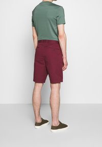 Pantaloni corti da uomo color bordeaux in morbido cotone misto, dotati di due tasche posteriori con bottoni e una vestibilità dritta classica, abbinati a una t-shirt verde.