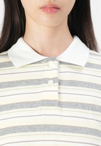 Striped polo shirt in yellow, gray, and beige, featuring a white collar, three buttons, and soft texture. "SAMSUG" logo on the fabric.
