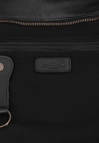 Sac en tissu noir avec deux fermetures éclair horizontales, un patch en cuir embossé "Gusti Leder studio" et une sangle en cuir avec un anneau en métal.