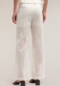 Witte, losvallende broek van geperforeerde stof met diamantpatronen, met een brede pijp en gedragen met zwarte hoge hakken.