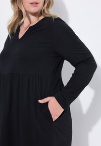 Robe noire à manches longues avec décolleté en V, taille cintrée et jupe froncée. Elle présente des poches latérales et une texture douce et lisse.