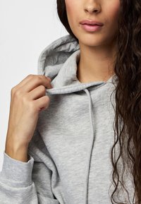 Pitkä kiharahiuksinen nainen pitää vaaleanharmaan hupun collaria, näyttäen kankaan tekstuurin ja kiristysnauhat.