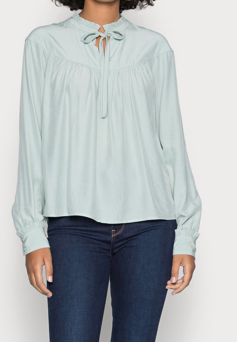 Blouse à manches longues bleu clair avec un col froncé et un détail à nouer. Tissu doux avec un design fluide et des poignets boutonnés. Associée à un pantalon en jean foncé.