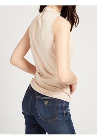 Mujer vistiendo una blusa beige sin mangas con cremallera en la espalda y jeans oscuros de Guess, posando con una mano en la cadera, dando la espalda a la cámara.