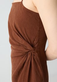 Abito senza maniche marrone realizzato in tessuto testurizzato, con un dettaglio a nodo attorcigliato sul lato per un interesse di design aggiuntivo.