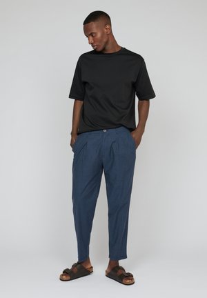 T-shirt nera a maniche corte, dalla vestibilità ampia. Pantaloni blu a pieghe, con gamba affusolata. Sandali marroni con fasce. Sfondo neutro.