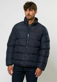 Marineblå pufferjakke med quiltet design, høy krage og rødt logo på brystet, med en glatt, blank overflate. Glidelås lukking.