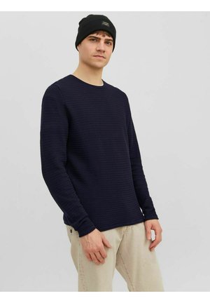 Navy textured sweater with long sleeves and a crew neckline, paired with light beige pants and a black knit beanie featuring a patch.