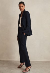 Žena stojící v tmavě modrém saku a kalhotách s bílým topem a černými páskovými vysokými podpatky na jednobarevném béžovém pozadí.