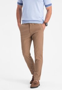 Mężczyzna ubrany w jasnoniebieską koszulę z krótkim rękawem, beżowe, dopasowane spodnie, brązowe mokasyny z frędzlami i brązowy zegarek na rękę, z rękami w kieszeniach.