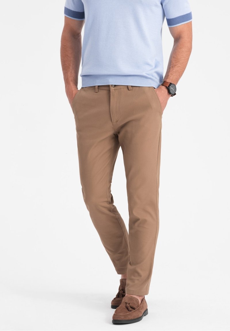 Mężczyzna ubrany w jasnoniebieską koszulę z krótkim rękawem, beżowe, dopasowane spodnie, brązowe mokasyny z frędzlami i brązowy zegarek na rękę, z rękami w kieszeniach.