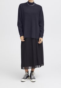 Pull en maille côtelée bleu marine avec col montant, associé à une jupe midi plissée noire et des baskets montantes noires avec semelles blanches.