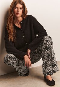 Cardigan noir texturé avec boutons, associé à un pantalon à carreaux noir et blanc à motif floral. Le modèle est accroupi, portant des chaussures plates noires, sur un fond neutre.
