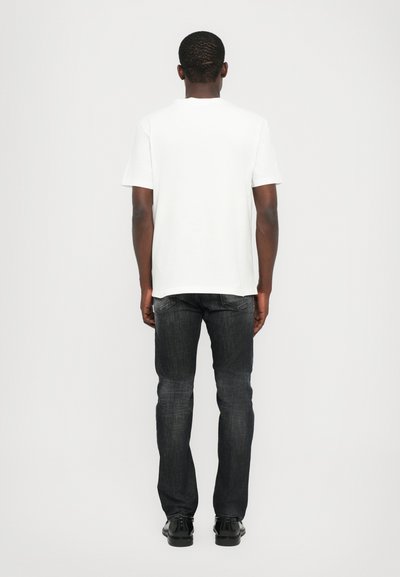 Camiseta de algodón blanca de manga corta, combinada con jeans gris oscuro y zapatos negros, vista desde la parte de atrás sobre un fondo liso.