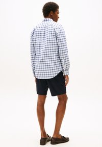 Le produit présente une chemise à manches longues à carreaux bleu et blanc avec des poignets à boutons, associée à un short bleu marine. Le tissu semble lisse et léger.