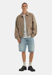 Giacca bomber marrone chiaro con zip frontale, caratterizzata da un colletto semplice e due tasche laterali, abbinata a pantaloni corti di jeans e scarpe nere.