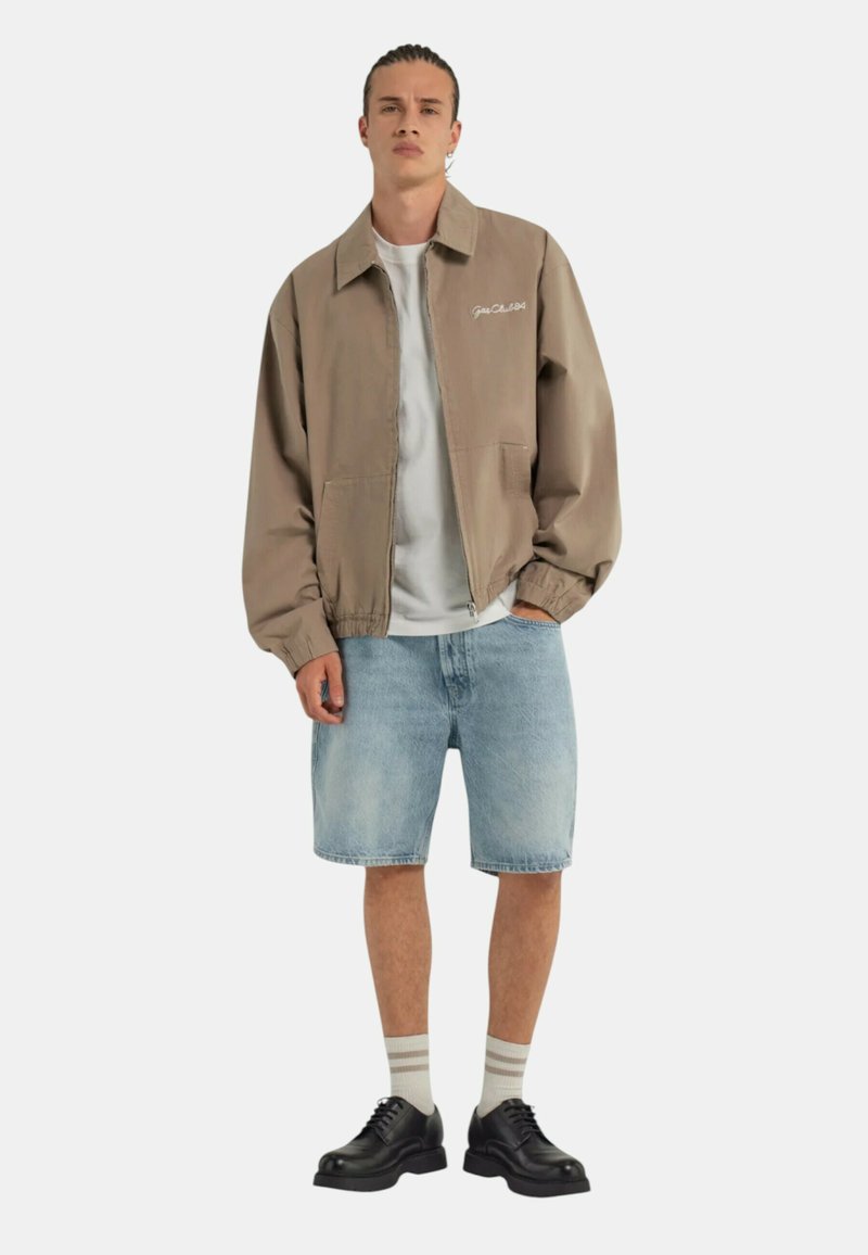 Giacca bomber marrone chiaro con zip frontale, caratterizzata da un colletto semplice e due tasche laterali, abbinata a pantaloni corti di jeans e scarpe nere.