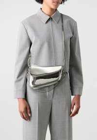 Borsa a tracolla metallica argento con pattina, texture liscia e tracolla regolabile, indossata sopra un outfit a scacchi bianco e nero.
