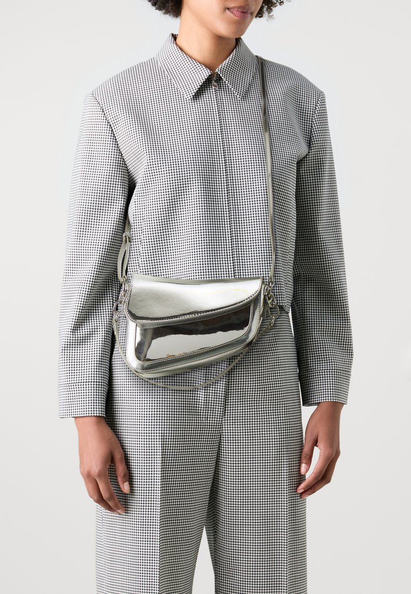 Borsa a tracolla metallica argento con pattina, texture liscia e tracolla regolabile, indossata sopra un outfit a scacchi bianco e nero.