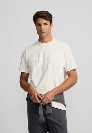 Hombre vestido con una camisa blanca de manga corta con texto en relieve, pantalones oscuros y un suéter gris atado alrededor de su cintura.