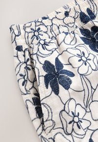 Weißer Stoff mit marineblauer Blumenstickerei, ausgestattet mit einem gerafften Bund und sichtbaren Nähdetails. Glatte Textur, lockere Passform.