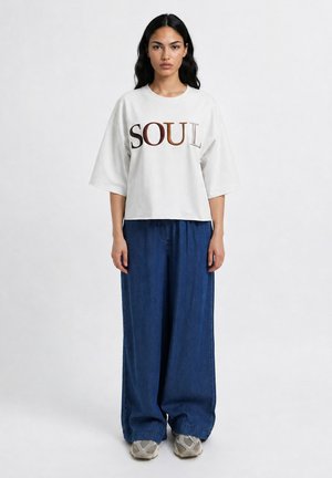 Woman with long dark hair stands facing forward wearing a white oversized t-shirt with "SOUL" text and wide blue jeans with sneakers.