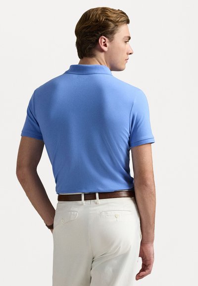 Ljust blå kortärmad polo t-shirt med en traditionell krage, bärs med krämfärgade byxor och ett brunt läderbälte, sedd bakifrån.