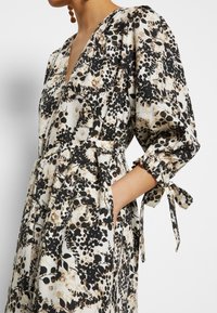 Kukkakuvioinen mekko, jossa on musta, beige ja valkoinen kuvio, V-pääntie, kolmanneksen hihat puffimuodossa sekä vyötärövyö solmuntarkoituksella.