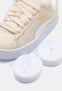 Sneaker en suède beige avec de larges lacets texturés, semelle en caoutchouc blanche avec un bord cranté, et deux lacets blancs enroulés à proximité. Détail du logo doré visible.