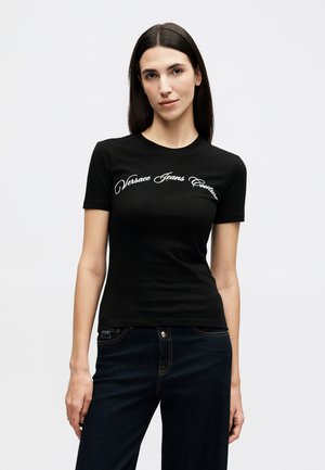 Femme aux longs cheveux noirs portant un t-shirt noir ajusté avec le logo "Versace Jeans Couture" et un jean foncé, debout devant un fond uni.