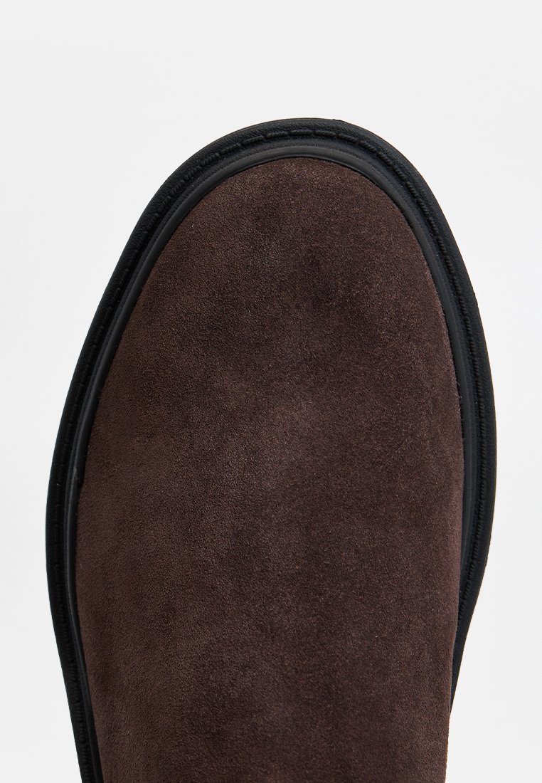 Bottines en daim marron avec un bout arrondi, dotées d'une semelle en caoutchouc noire et de détails de couture le long du bord. Texture lisse.