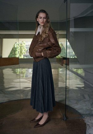 Chaqueta bomber de cuero marrón con cuello de piel, combinada con una falda plisada gris oscuro y zapatos planos marrones de punta afilada.