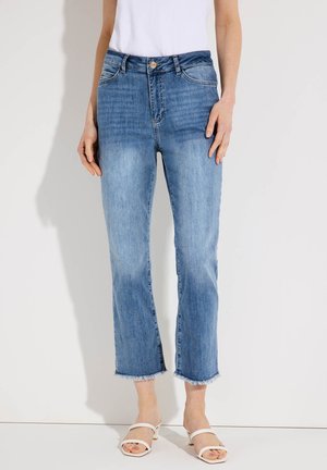 Frau trägt hellblaue, hochgeschnittene, kurz geschnittene Jeans mit ausgefransten Säumen und weißen Sandalen mit Absatz, steht vor einem schlichten weißen Hintergrund.