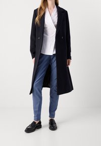 Frau, die einen langen schwarzen Mantel, eine weiße Wickelbluse, maßgeschneiderte blaue Hose und schwarze Mokassins vor einem schlichten weißen Hintergrund trägt.