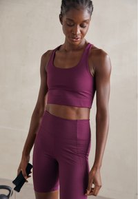 Aktīvās sporta apģērba komplekts tumši purpursarkanā krāsā, sastāv no pieguloša krūštura un augsta vidukļa šortiem. Gluda tekstūra ar minimālām šuvēm un sportisku dizainu.