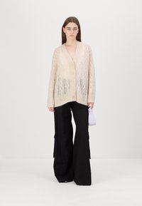 Cardigan tricotat de culoare crem, cu un model texturat, guler în V și cinci nasturi frontali, asortat cu pantaloni largi negri.