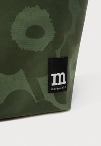 Tumši zaļš audums ar abstraktu ziedu rakstu un melnu etiķeti ar baltu "m" un "marimekko" tekstu, uzšūtu netālu no apakšējās malas.