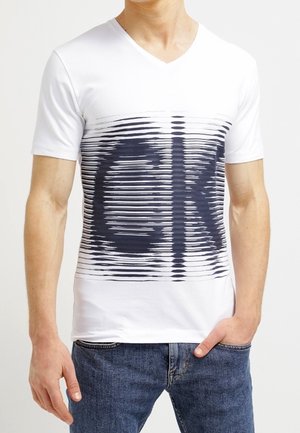 Man in een witte V-hals T-shirt met een vervormd zwart "CK" logo, gecombineerd met blauwe denim jeans, staande met één hand in de zak.