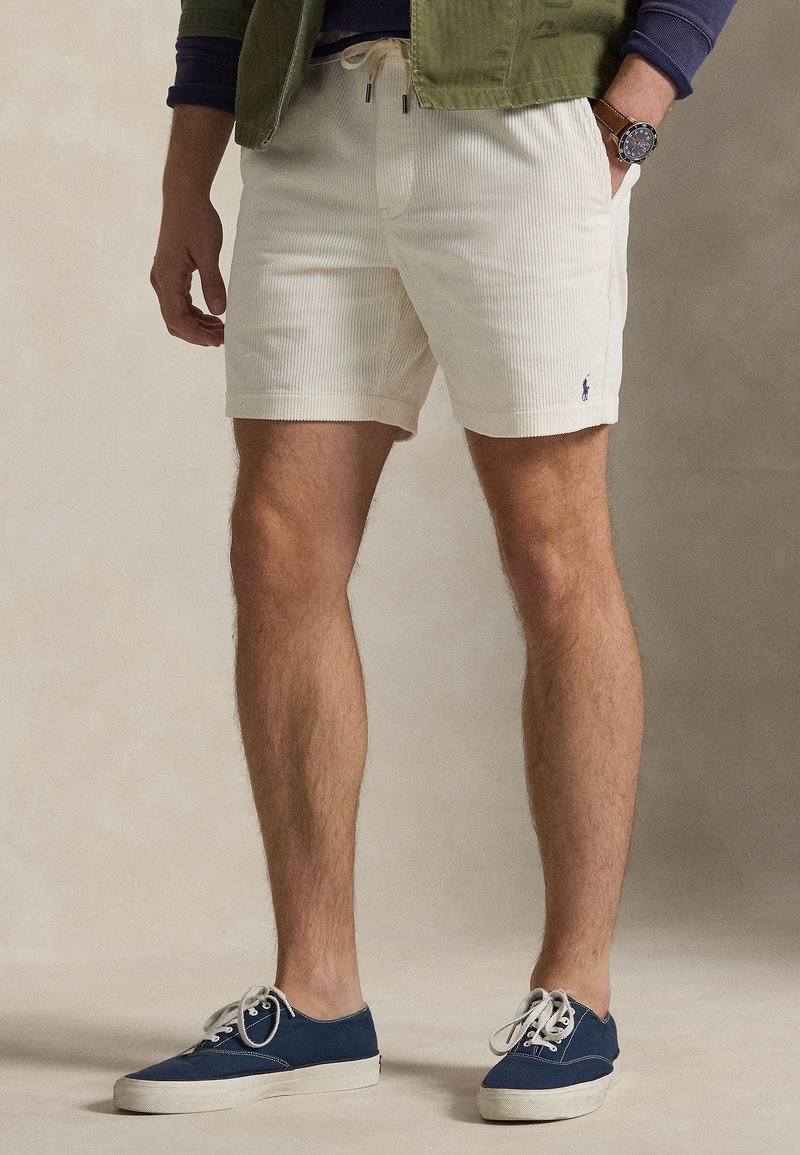 Homme portant un short en velours côtelé blanc, des baskets bleu marine, une veste verte et une montre-bracelet, debout avec une main dans la poche.