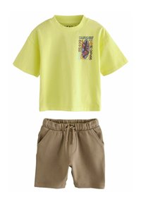 Gul bomullströja med grafiskt tryck och korta ärmar, ihop med bruna shorts med dragsko i mjukt material.