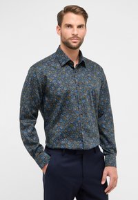 Man in een donkerblauwe bloemenpatroon lange mouwen shirt met marineblauwe broek, staande met één hand in de zak tegen een neutrale achtergrond.