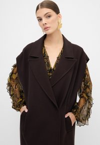 Brązowy, oversize'owy płaszcz z kołnierzem i przednimi kieszeniami. Pod nim bluzka w kwiatowy wzór z falbaniastymi, przezroczystymi rękawami. Widoczne złote kolczyki.