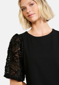Czarna sukienka z krótkimi, teksturowanymi przezroczystymi rękawami z falbaniastymi poziomymi pasami. Gładkie body, okrągły dekolt, elegancki, ogólny design.