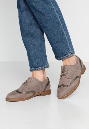 Mocassins en daim taupe avec des coutures décoratives et un accent contrastant marron foncé. Bout rond, design à lacets et semelle plate beige.