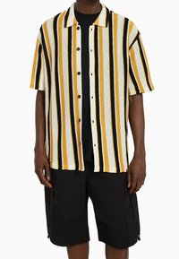 Koszula z krótkim rękawem w pionowe paski w kolorze żółtym, czarnym i białym, z zapięciem na guziki z przodu i kołnierzykiem rozłożonym. Połączona z czarnymi szortami.