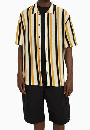 Camisa de manga corta con rayas verticales en amarillo, negro y blanco, que presenta un cierre frontal con botones y un cuello amplio. Combinada con pantalones cortos negros.